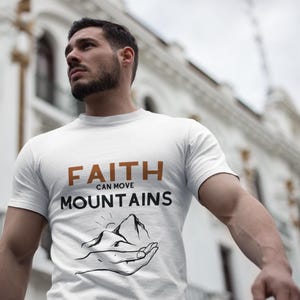 Pode incluir: Camiseta branca com a frase "FAITH CAN MOVE MOUNTAINS" em castanho e preto. O design inclui uma cordilheira e uma mão. A camiseta é feita de um material macio.