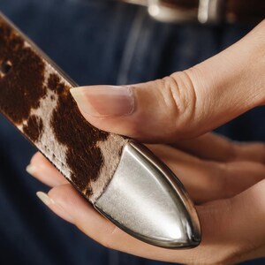 May include: A belt strap featuring a brown and white cowhide pattern with a smooth, pointed silver-toned metal tip. The strap has visible stitching along the edges and a series of punched holes for adjustment, showcasing a textured finish.