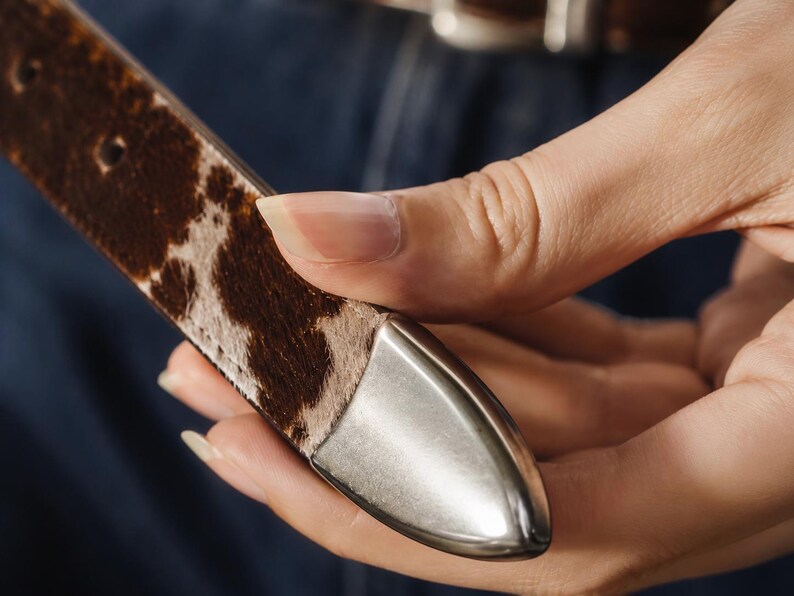 May include: A belt strap featuring a brown and white cowhide pattern with a smooth, pointed silver-toned metal tip. The strap has visible stitching along the edges and a series of punched holes for adjustment, showcasing a textured finish.