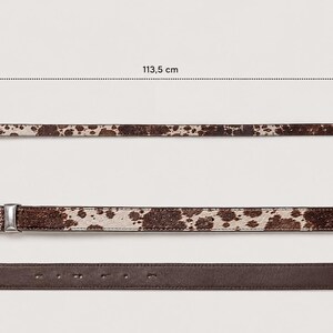 May include: A brown and white cow print belt with a silver-toned metal buckle. The belt measures 113.5 cm in total length and 1.9 cm in width. The underside of the belt is a solid dark brown leather material with visible adjustment holes.