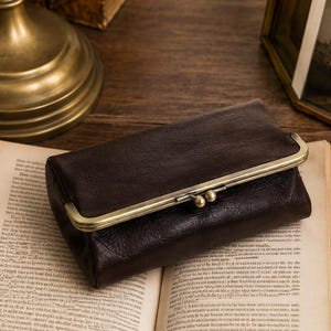 May include: A dark brown rectangular wallet with a textured finish and a metallic gold-toned frame closure. The wallet features a hinged clasp with two small spherical knobs at the top, providing a secure opening mechanism for the main compartment.