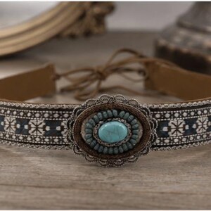 Peut inclure: Un collier ras-du-cou en cuir marron avec un médaillon central ovale en métal argenté orné d'une pierre turquoise entourée de petites perles bleues. La bande présente un motif tissé bleu foncé et blanc avec des liens de serrage en cuir.