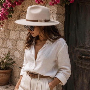 May include: A beige fedora hat with a brown leather band and a gold-toned metal buckle, paired with a white long-sleeved button-down shirt, a brown leather belt, and dark-tinted sunglasses. The outfit features neutral tones and classic textures.