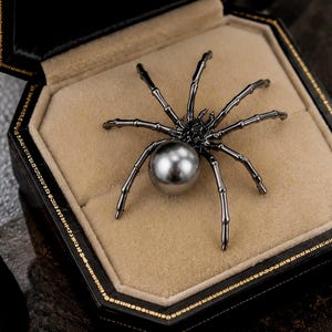 May include: A spider-shaped brooch featuring a dark metallic body with eight segmented legs and a large, smooth, spherical grey pearl serving as the abdomen, presented inside a black octagonal jewelry box with a beige velvet interior lining.