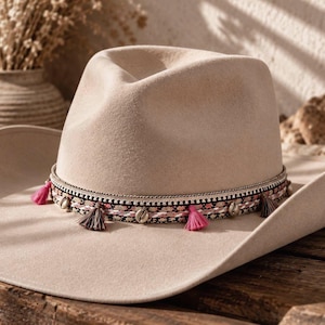 May include: A beige felt cowboy hat featuring a wide brim and a pinched crown. The hat is adorned with a decorative band that includes small pink tassels, dark grey tassels, and metallic accents arranged in a repeating pattern around the base.