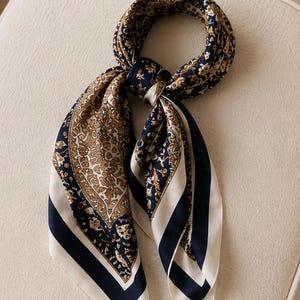 May include: A square scarf featuring a dark navy blue and cream colour palette with an intricate gold and brown paisley pattern. The scarf is tied in a knot and displays a wide solid navy blue border along the edges with a cream-coloured trim.