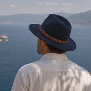 Op de afbeelding: Een marineblauwe fedora hoed voorzien van een bruine leren band met een kleine metalen gesp. De hoed heeft een gestructureerde kroon en een brede rand, wat zorgt voor een klassieke uitstraling. Het materiaal oogt glad en uniform.