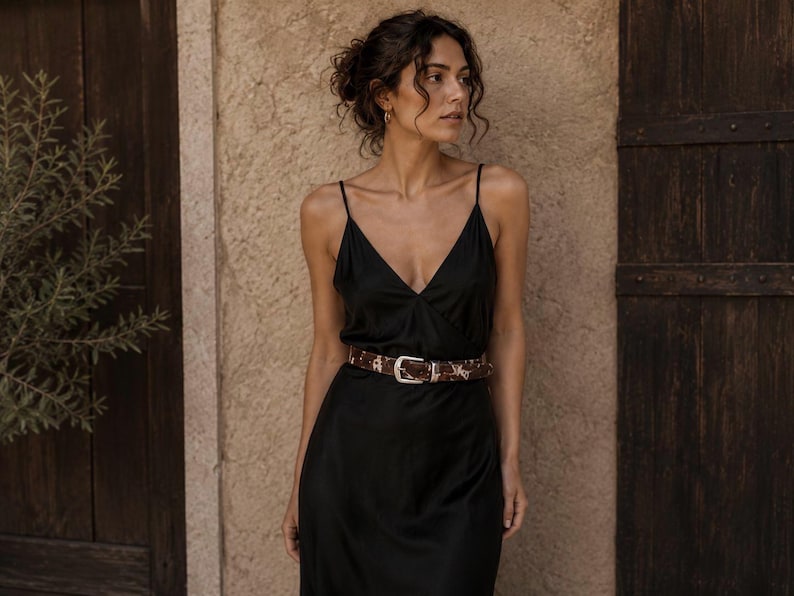 May include: A black midi-length slip dress with thin spaghetti straps and a V-neckline, cinched at the waist with a brown and white patterned belt featuring a silver-toned metal buckle. The dress is made of a smooth, satin-like fabric.
