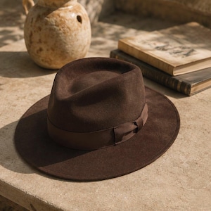 Op de afbeelding: Een donkerbruine vilten fedora-hoed met een gestructureerde kroon en een brede, platte rand. De hoed is voorzien van een bijpassende donkerbruine grosgrain lintband die aan de zijkant tot een kleine strik is geknoopt voor een strakke uitstraling.
