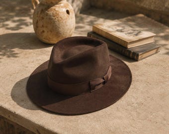 Op de afbeelding: Een donkerbruine vilten fedora-hoed met een gestructureerde kroon en een brede, platte rand. De hoed is voorzien van een bijpassende donkerbruine grosgrain lintband die aan de zijkant tot een kleine strik is geknoopt voor een strakke uitstraling.