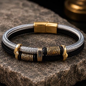 Puede incluir: Una pulsera de doble hebra que presenta un cordón trenzado de tono plateado y un cordón negro. La pieza incluye detalles de metal en tono dorado, incluyendo dos cuentas cilíndricas con patrones grabados intrincados, un espaciador central liso y un cierre.