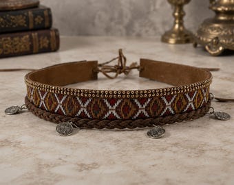 Op de afbeelding: Een bruine choker-halsketting met een geweven band in een geometrisch patroon van rood, geel en wit, geaccentueerd door een rij kleine goudkleurige studs. Het sieraad bevat een gevlochten bruine koordlaag en diverse bungelende zilverkleurige muntbedels.