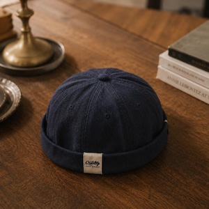 May include: A navy blue brimless cap featuring a rounded crown with six panels and a small fabric button at the top. A rectangular beige patch is centered on the rolled cuff, displaying the word Quality in a dark script font with smaller text below.