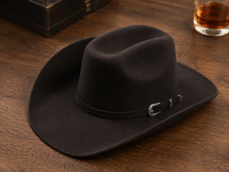 Op de afbeelding: Een donkerbruine vilten cowboyhoed met een ingedeukte kroon en een brede, gebogen rand. De hoed is voorzien van een dunne, bijpassende donkerbruine hoedband met aan de zijkant een decoratieve, gegraveerde zilverkleurige metalen gesp.