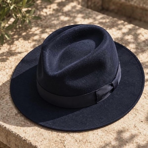 May include: A dark navy blue felt fedora hat featuring a structured crown with a centre crease and a wide, flat brim. The hat is accented with a matching dark navy blue satin ribbon band that encircles the base of the crown.