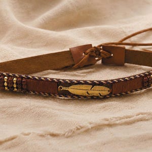 Op de afbeelding: Een bruine leren riem met een centrale veerbedel van goudkleurig metaal, geaccentueerd met donkerbruine houten kralen en dunne gevlochten koorddetails. De riem heeft een striksluiting met lange, smalle leren banden voor verstelling.