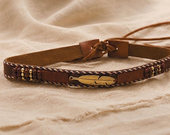 Op de afbeelding: Een bruine leren riem met een centrale veerbedel van goudkleurig metaal, geaccentueerd met donkerbruine houten kralen en dunne gevlochten koorddetails. De riem heeft een striksluiting met lange, smalle leren banden voor verstelling.