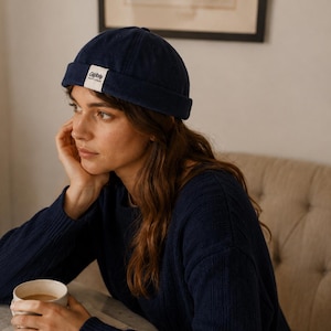 Peut inclure: Un bonnet docker bleu marine avec un bord roulé et un écusson rectangulaire blanc sur le devant. L'écusson porte l'inscription "Gleby" en lettres sombres. Le bonnet est fabriqué dans un tissu texturé et se porte ajusté sur la tête.