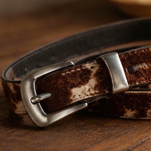May include: A belt featuring a brown and white cowhide pattern with a textured, hairy surface. The belt includes a rectangular, brushed silver-toned metal buckle and a matching silver-toned metal keeper loop, secured with a small dark metal rivet.