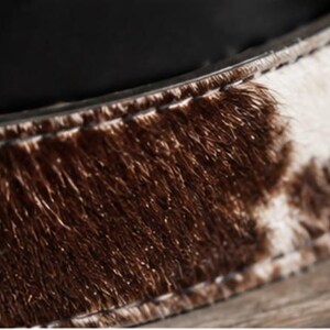 May include: A belt featuring a textured cowhide pattern with patches of dark brown and white fur. The strap has visible stitching along the edges and is lined with a smooth black material on the interior side, showing a clear contrast in textures.