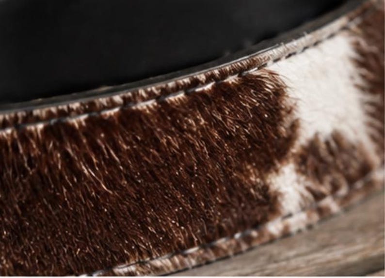May include: A belt featuring a textured cowhide pattern with patches of dark brown and white fur. The strap has visible stitching along the edges and is lined with a smooth black material on the interior side, showing a clear contrast in textures.