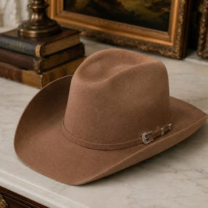 Op de afbeelding: Een beige vilten cowboyhoed met een gestructureerde kroon en een brede, gebogen rand. De hoed is voorzien van een bijpassende hoedband versierd met een kleine, gedetailleerde zilverkleurige metalen gesp met gravures en heldere steentjes.