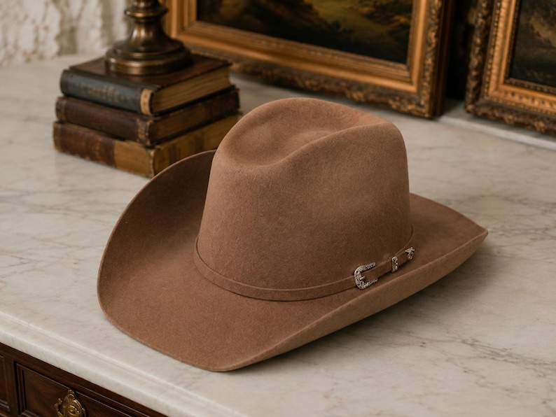 Op de afbeelding: Een beige vilten cowboyhoed met een gestructureerde kroon en een brede, gebogen rand. De hoed is voorzien van een bijpassende hoedband versierd met een kleine, gedetailleerde zilverkleurige metalen gesp met gravures en heldere steentjes.