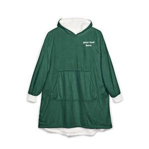 Pode incluir: Uma camisola com capuz oversized verde-escura com forro interior branco, mangas compridas com punhos brancos e um bolso canguru frontal grande. O texto "your text here" está impresso a branco na parte superior esquerda do peito.