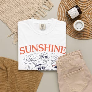Può includere: Una maglietta bianca con la scritta "SUNSHINE" in arancione e una grafica a tema spiaggia. La maglietta è abbinata a pantaloni beige, un tappetino intrecciato e una candela. La maglietta è in materiale Comfort Colors.