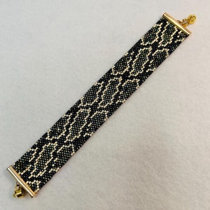 May include: A beaded bracelet with a snake scale pattern in black, green, and cream colors. The bracelet has gold-colored metal end caps and a clasp. The bracelet is approximately 1 inch wide.