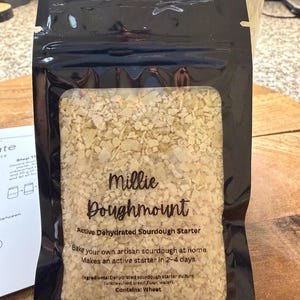 May include: A black package of Millie Doughmount active dehydrated sourdough starter. The package has a clear window showing the contents, which are light beige flakes. Text on the package says to bake artisan sourdough at home and makes an active starter in 2-4 days. Net weight is 20g.