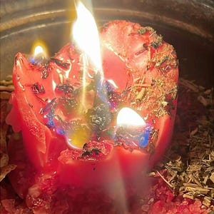 May include: A red pillar candle burning with multiple flames, embedded with dried herbs and botanical pieces. The melting wax pools around the base, which is surrounded by coarse red salt crystals and scattered dried plant matter in a metal container.
