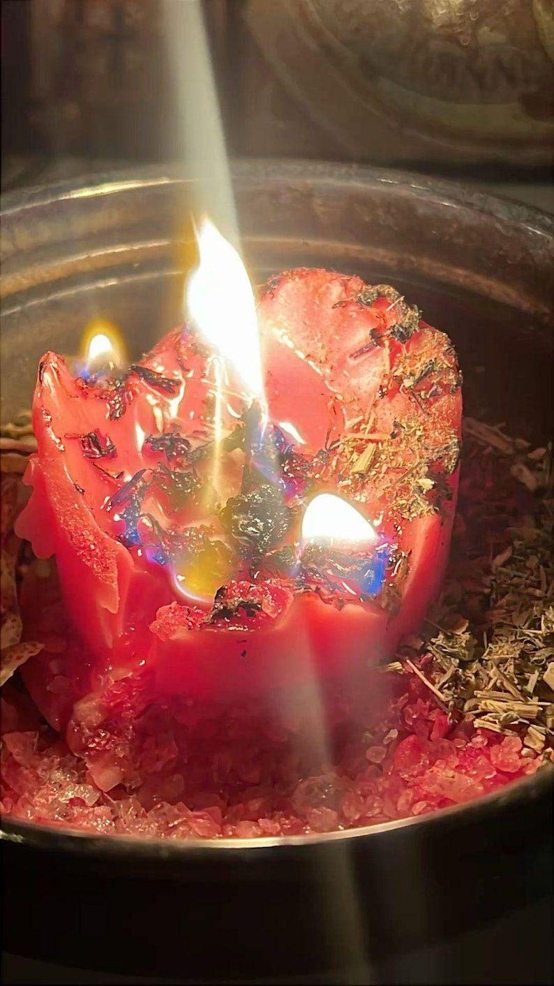 May include: A red pillar candle burning with multiple flames, embedded with dried herbs and botanical pieces. The melting wax pools around the base, which is surrounded by coarse red salt crystals and scattered dried plant matter in a metal container.