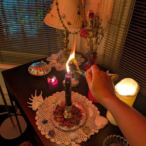 May include: A dark purple candle sits in a silver-toned holder on a white lace doily, surrounded by various seashells and crystals. A hand holds a pair of metal tweezers to light a small piece of paper in the candle flame, with a lamp nearby.