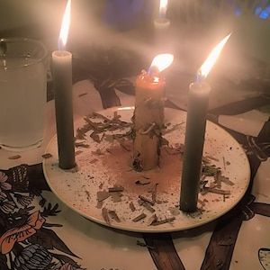 May include: A light-colored ceramic plate holds a central thick candle surrounded by three slender dark gray candles, all lit with visible flames. The plate is scattered with dried herbs and sits on a surface featuring a floral and botanical illustration.