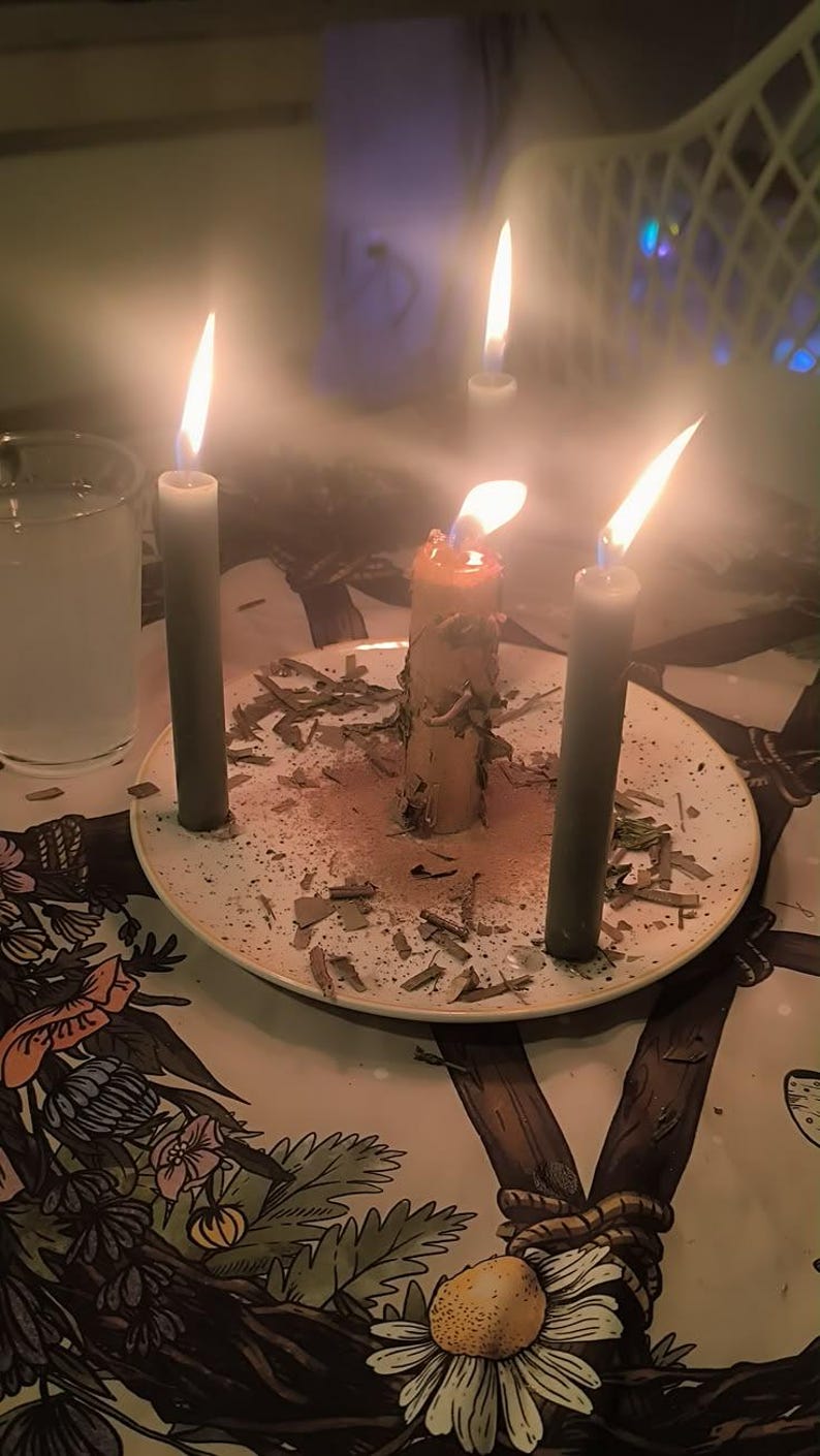 May include: A light-colored ceramic plate holds a central thick candle surrounded by three slender dark gray candles, all lit with visible flames. The plate is scattered with dried herbs and sits on a surface featuring a floral and botanical illustration.