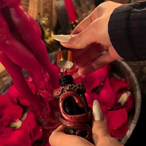May include: A small black glass dropper bottle featuring a metallic red snake coiled around the neck and body. The bottle has a gold-colored dropper cap and is being held over a collection of red rose petals near a red candle.