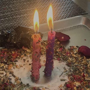 May include: Two lit taper candles, one pink and one purple, stand upright on a silver tray. The candles are embedded with dried flower petals and herbs. A ring of loose dried botanicals and rose petals surrounds the base of the candles.