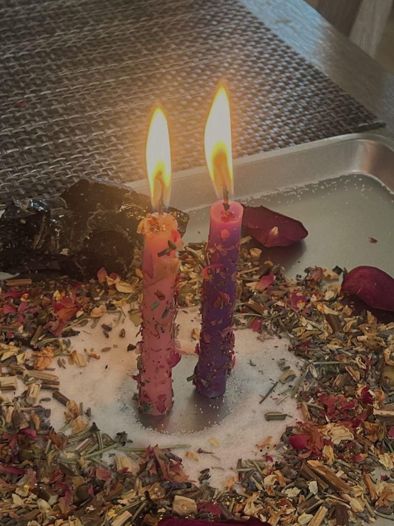 May include: Two lit taper candles, one pink and one purple, stand upright on a silver tray. The candles are embedded with dried flower petals and herbs. A ring of loose dried botanicals and rose petals surrounds the base of the candles.