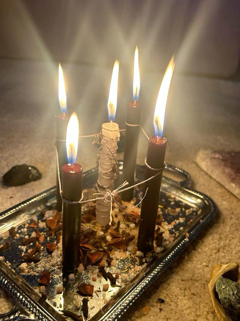 May include: Four dark brown candles and one central white candle, all lit with bright flames, are arranged on a rectangular silver tray. The candles are connected by thin twine. The tray is scattered with dried herbs, spices, and salt.