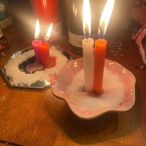May include: Two lit candles, one white and one orange, stand upright in a pink scalloped dish filled with white salt. Beside them, two smaller lit candles, one pink and one orange, sit on a hexagonal mirror tray also covered in white salt.
