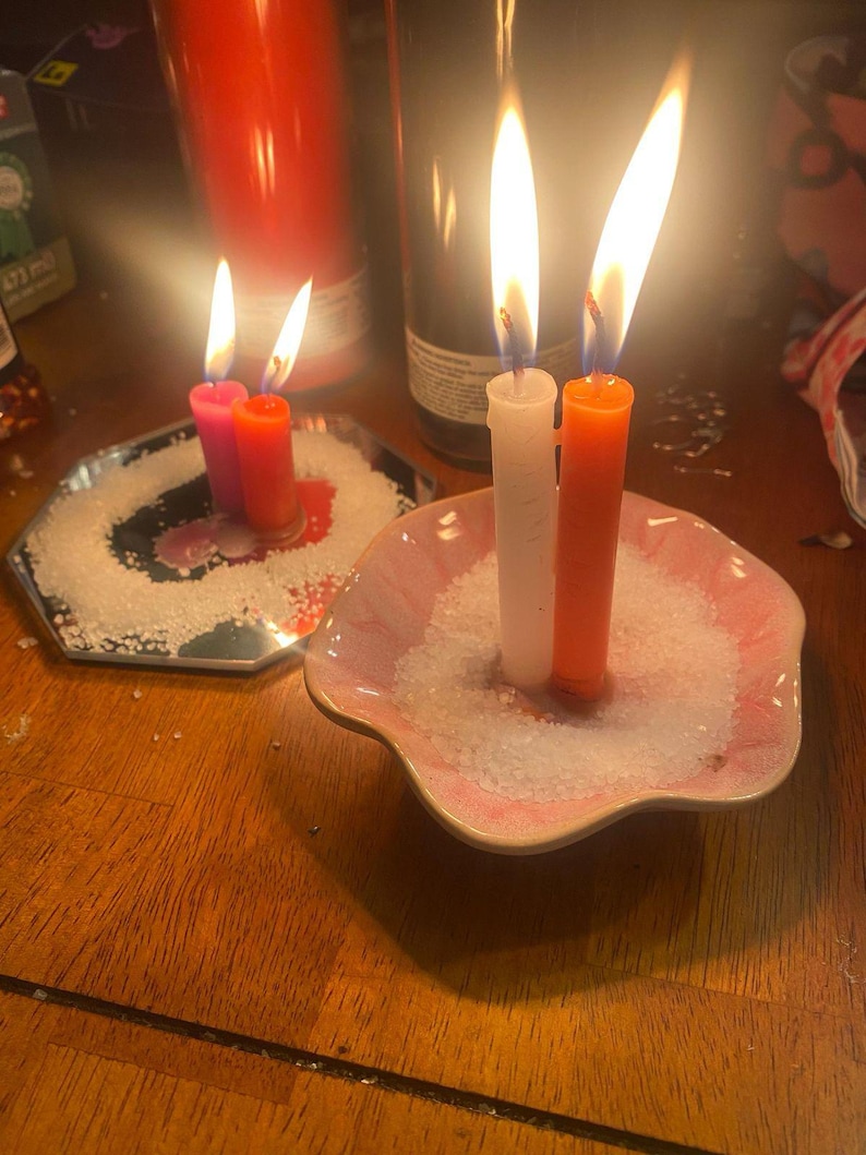 May include: Two lit candles, one white and one orange, stand upright in a pink scalloped dish filled with white salt. Beside them, two smaller lit candles, one pink and one orange, sit on a hexagonal mirror tray also covered in white salt.