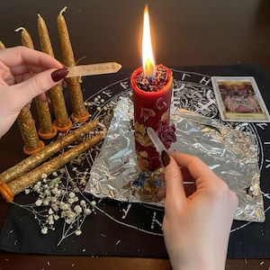 Pode incluir: Uma vela pilar vermelha alta com chama acesa, decorada com pétalas de flores secas e uma ponta de cristal transparente. Várias velas cónicas douradas estão próximas e uma carta de tarot repousa sobre um pano preto com desenho circular.