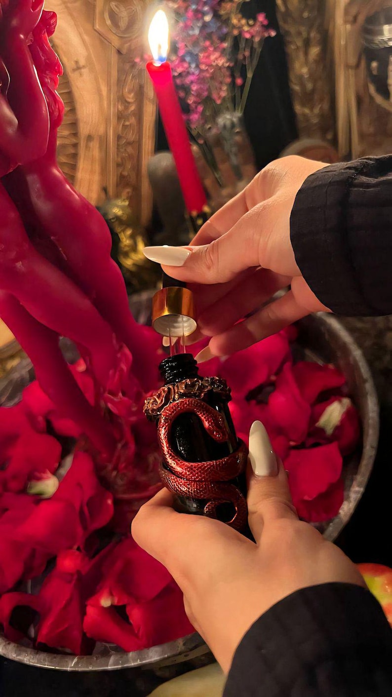 May include: A small black glass dropper bottle featuring a metallic red snake coiled around the neck and body. The bottle has a gold-colored dropper cap and is being held over a collection of red rose petals near a red candle.