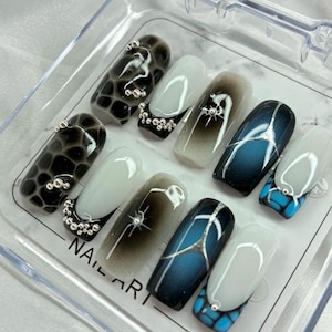 Peut inclure: Un ensemble de dix faux ongles dans un boîtier en plastique transparent marqué NAIL ART. Les motifs incluent des textures peau de reptile noires et bleues, des manucures françaises blanches avec des perles argentées et des étoiles argentées.