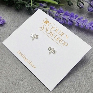 May include: Sterling silver dragonfly stud earrings on a white card with the Golden Snowdrop logo. The earrings are small and detailed, with a dragonfly design. The card is on a grey surface with purple flowers in the background.