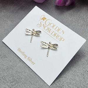 May include: A pair of sterling silver dragonfly stud earrings. The earrings are displayed on a white card with the text "Golden Snowdrop" and "Sterling Silver". The earrings are silver and have detailed wings and bodies.