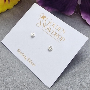 May include: A pair of sterling silver stud earrings with a spiral design, displayed on a white card. The card features the text "Golden Snowdrop" and "Sterling Silver". The earrings are small and minimalist.