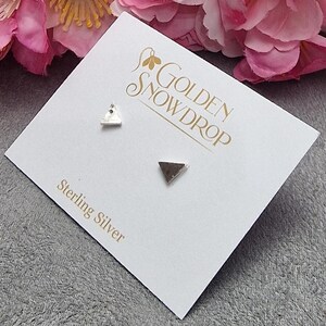 May include: Sterling silver triangle stud earrings displayed on a white card with the text "Golden Snowdrop". The card is on a gray surface, with pink flowers in the background. The earrings are small, geometric, and minimalist.