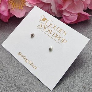 May include: A pair of sterling silver stud earrings with a smooth, oval design. The earrings are displayed on a white card with the text "Golden Snowdrop" and "Sterling Silver" in gold lettering. Pink flowers are in the background.